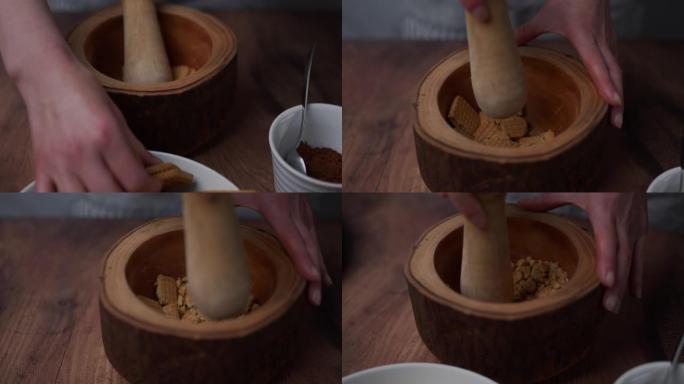 一个女人的手从一个装有饼干的白色碗中取出几块饼干，然后将它们放在木杵和研钵中，将其粉碎并磨成细粉。