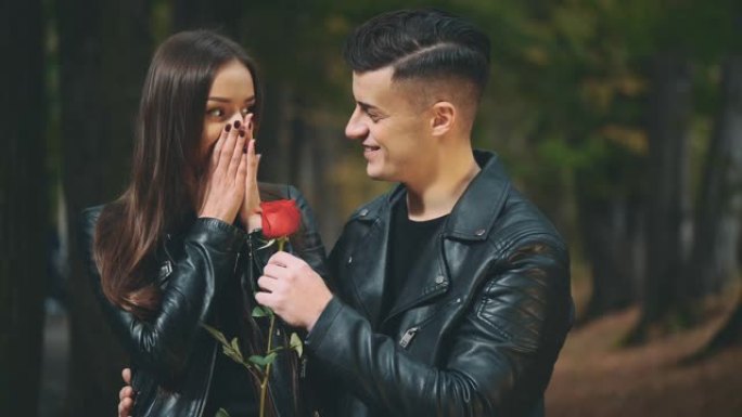 穿着黑色皮大衣的黑发女孩正在等她的男朋友。全黑的男孩闭上了女友的眼睛，献上一朵红玫瑰。背景模糊。复制