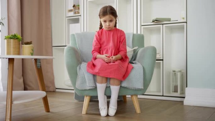 来自压力和沮丧的粉红色小女孩独自坐在家里的扶手椅上，悲伤地往下看