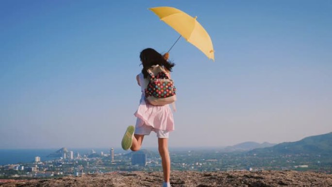 女孩背着背包，拿着黄色的伞在城市景观的顶部跳舞，生活方式的概念。