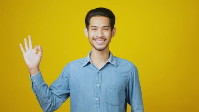 年轻的亚洲男子微笑着展示他的好兆头，快乐的亚洲男子看着相机，同时在工作室里站在黄色背景上展示好的手势