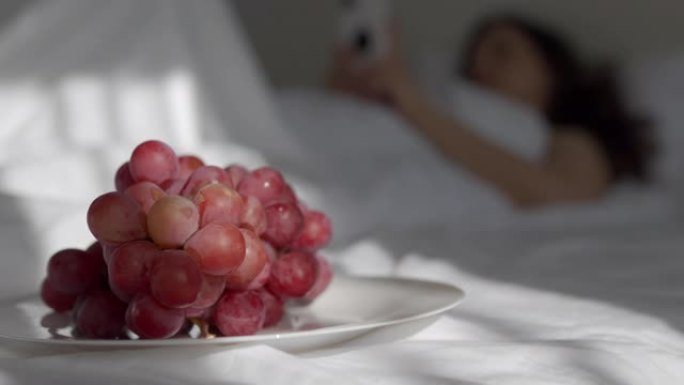 女人躺在床上拿着手机，阳光落在红葡萄盘子上