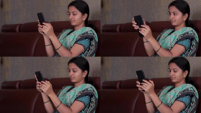 穿着便服的女孩坐在家里的沙发上忙碌在手机上-印度妇女在家中使用社交媒体，互联网，数字鸿沟和技术的概念