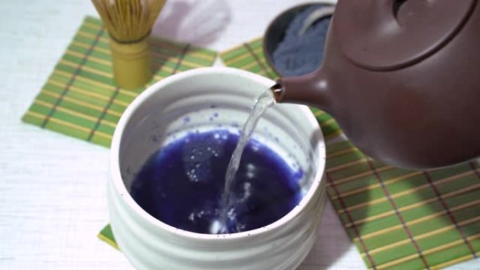 在白碗中加入热水，上面装有蓝色抹茶茶粉，竹制tusaku放在板上。