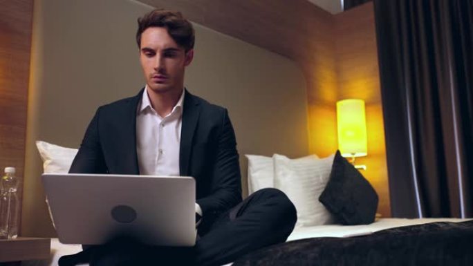年轻的认真商人坐在床上，在酒店房间里使用笔记本电脑