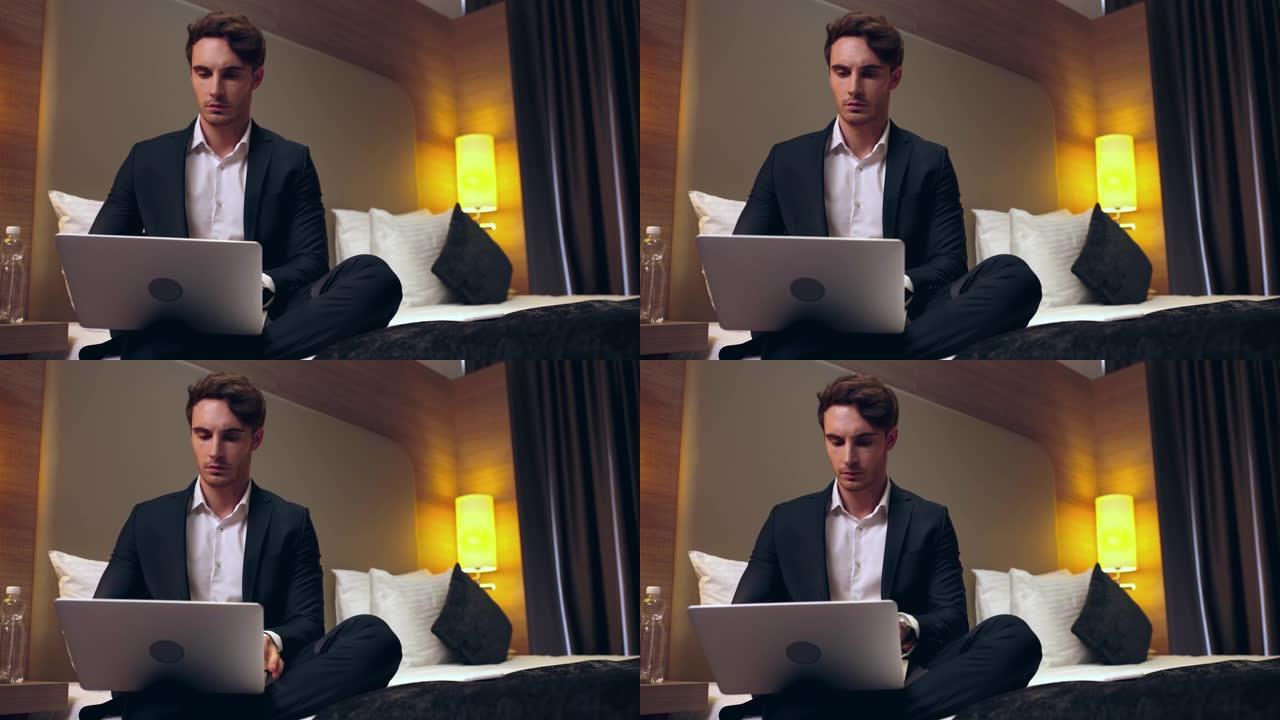 年轻的认真商人坐在床上，在酒店房间里使用笔记本电脑