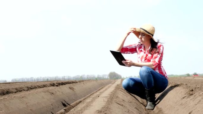 女农民，农艺师坐在田间特殊的土壤行之间。她使用片剂测试了中耕者种植马铃薯的质量。现代农业马铃薯种植。