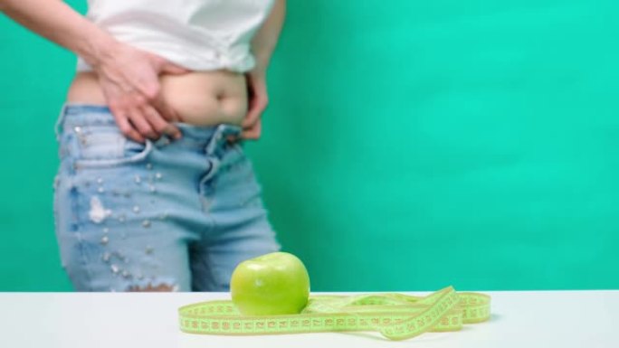 一个女人在卷尺和青苹果的背景下，触摸并测量她的油腻的胃，体重检查，腹部周围的衣服