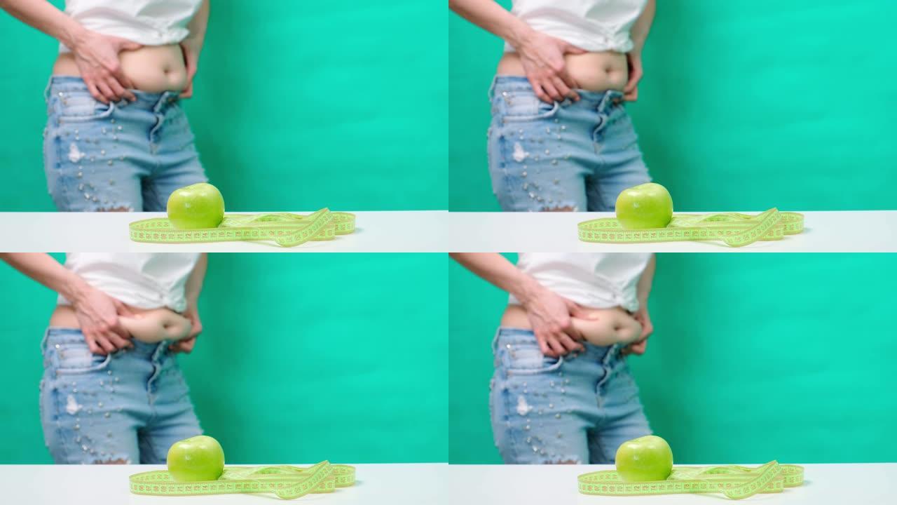 一个女人在卷尺和青苹果的背景下，触摸并测量她的油腻的胃，体重检查，腹部周围的衣服