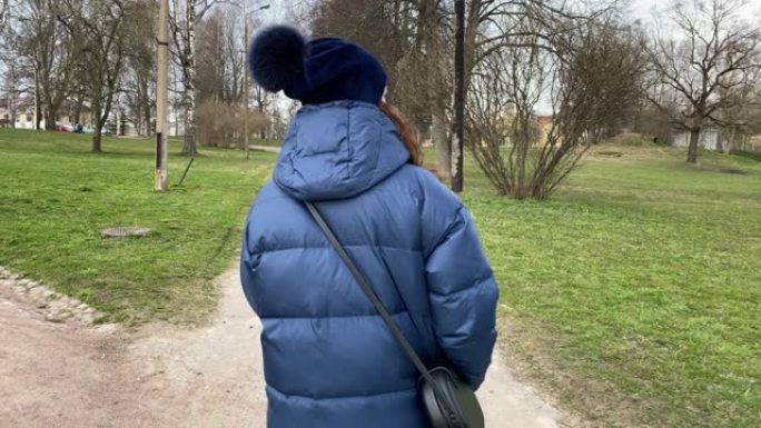 初春穿着冬季夹克走在公园人行道上的女人的后视图