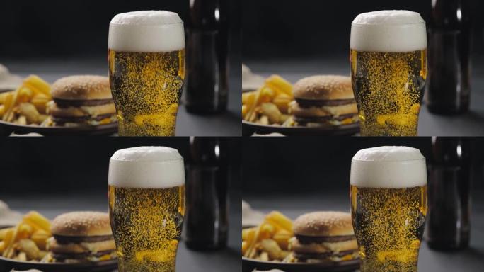 在玻璃杯中发泡啤酒，气泡冒出，顶部有白色泡沫。背景模糊，炸薯条和汉堡放在黑盘上。快餐。特写