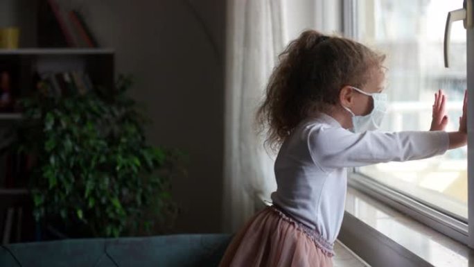 新型冠状病毒肺炎冠状病毒的概念，小女孩戴着面具透过窗户看，检疫在家里。检疫期间因COVID而悲伤的孩