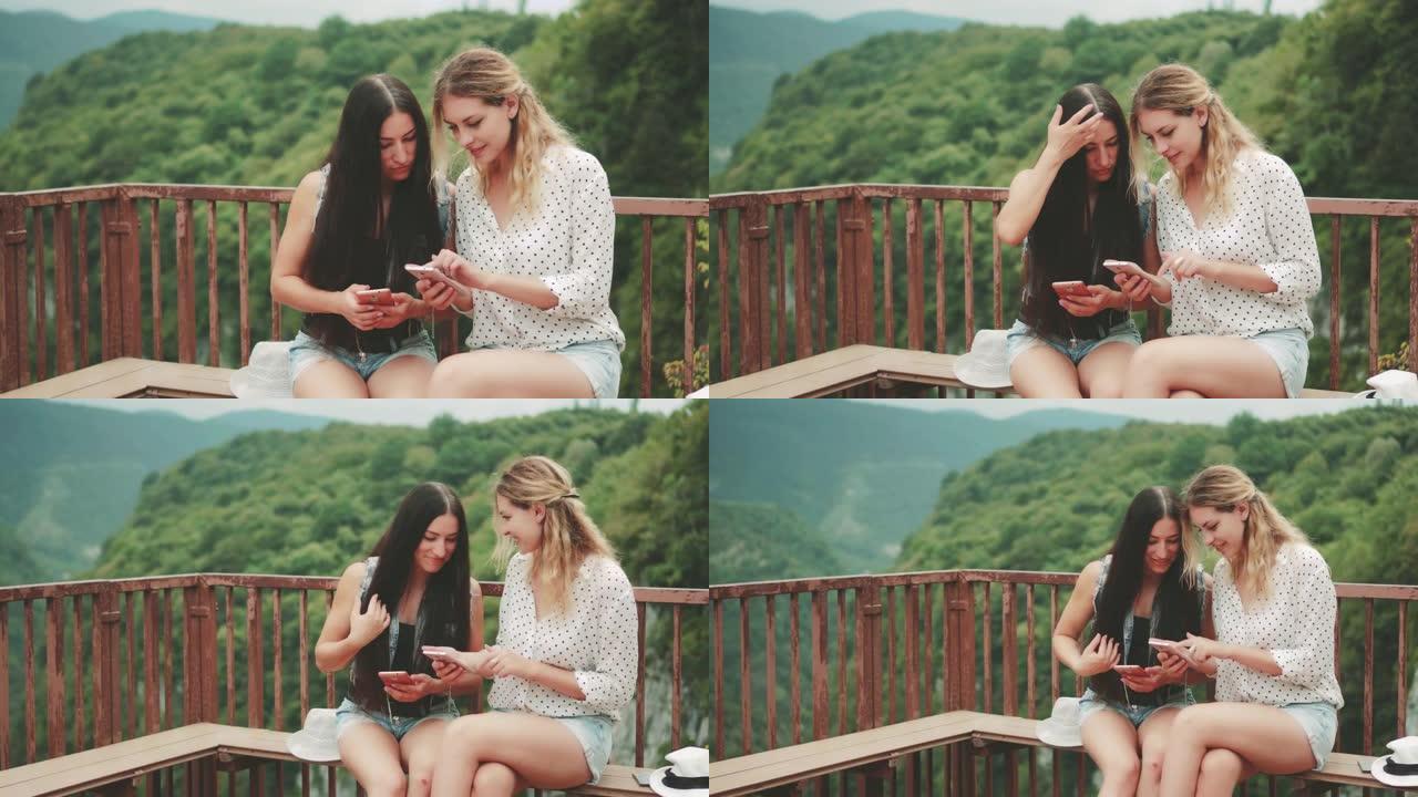 两个迷人的女人坐在上面的长椅上，山景如画。女孩在手机上显示照片朋友。依赖现代小玩意。快乐旅游乔治亚O
