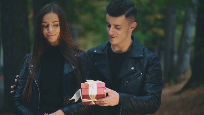 穿着黑色皮大衣的黑发女孩正在等她的男朋友。全黑的男孩正在给他可爱的女朋友一个红色包装的礼物盒子。背景