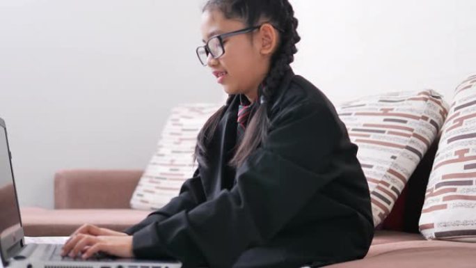 一个戴着眼镜，穿着黑色西装的亚洲小女孩，一个坐在棕色沙发上的女商人正在幸福地在笔记本电脑上打字。
