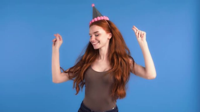 穿着党帽和卡其色衬衫的姜模特。她在蓝色背景上摆姿势时跳舞，微笑和拍手。生日快乐概念。特写，慢动作