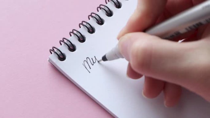在白色记事本纸上写下我的亲爱的。粉红色背景上的女性手握笔笔记或字母，特写