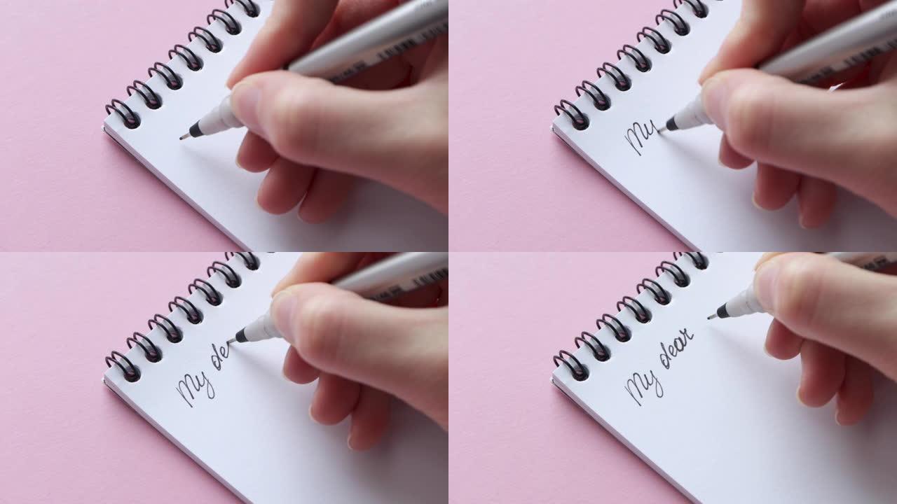 在白色记事本纸上写下我的亲爱的。粉红色背景上的女性手握笔笔记或字母，特写