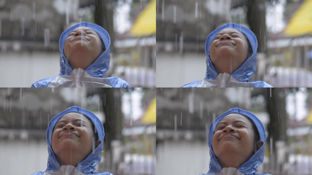 穿着蓝色雨衣的快乐年轻亚洲女孩站在屋前的雨中微笑着。
