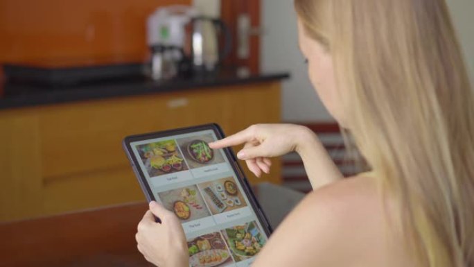 年轻女子用平板电脑在网上订购午餐食物。她坐在公寓的厨房里