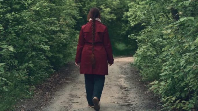 穿着红色夹克的年轻女子沿着绿色春天森林的小路散步，后视