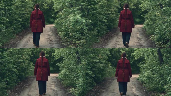 穿着红色夹克的年轻女子沿着绿色春天森林的小路散步，后视