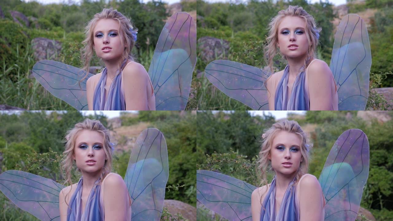 穿着蓝色连衣裙和透明翅膀的美丽年轻女孩站在草地上，看着相机。穿着童话公主服装的女孩。