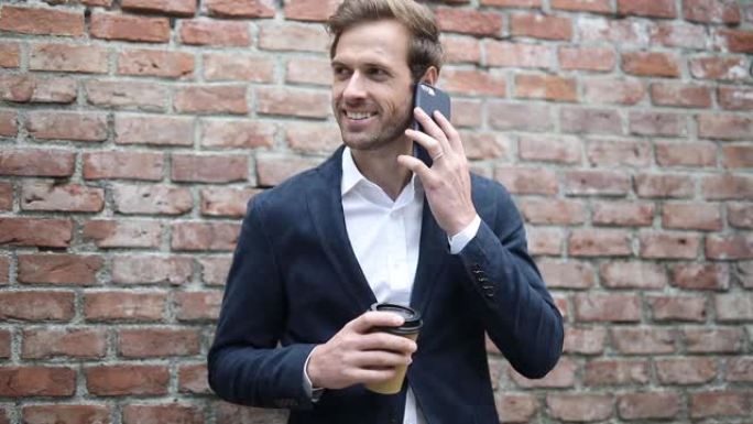 金发英俊的商人靠在砖墙上，喝一杯咖啡，掏出手机然后说话