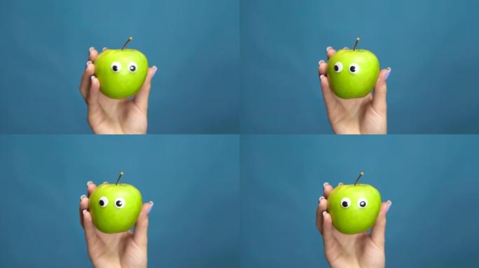绿苹果与眼睛在一个女人的手特写。苹果在蓝色背景上摇晃和扭曲眼睛。慢动作。