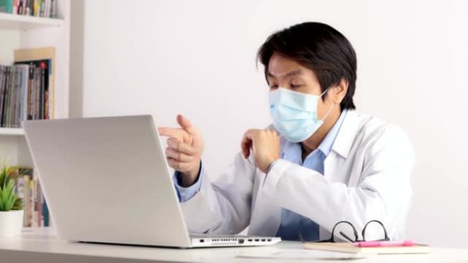 亚洲男性医生戴着口罩，通过视频会议系统与笔记本电脑一起工作