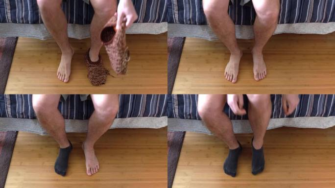 一个白人男性模特的特写镜头，坐在床边，从他破旧的棕色针织拖鞋中脱下，穿上黑色的脚踝袜子，然后走开。