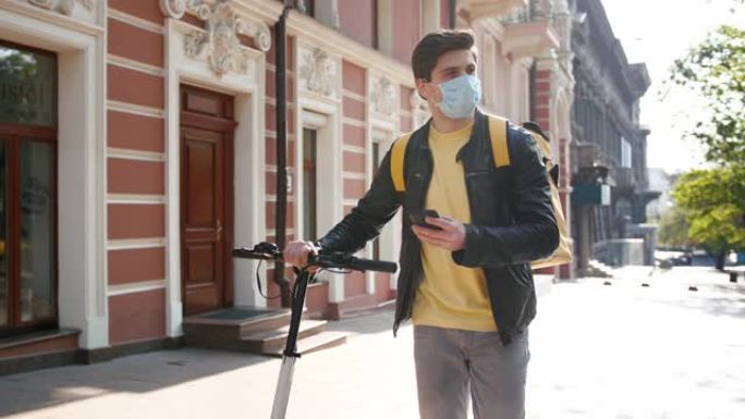 送货员带黄色袋装和防护面罩，带着电动滑板车行走，在城市中运送食物，并使用智能手机
