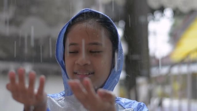 穿着蓝色雨衣的可爱的年轻亚洲女孩喜欢在房子前玩雨。