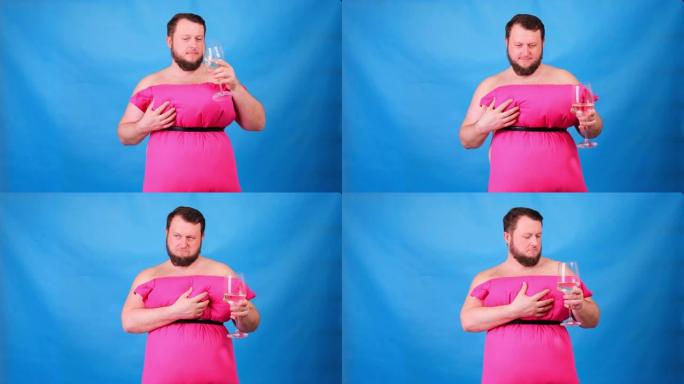 穿着粉红色枕头连衣裙的有趣的大胡子家伙从蓝色背景的玻璃杯中喝葡萄酒。疯狂的检疫。时尚2020。放在枕
