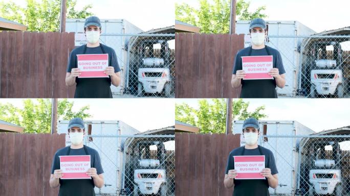 年轻的仓库工人举着 “停业” 的牌子，戴着口罩在他的工作场所关闭的入口