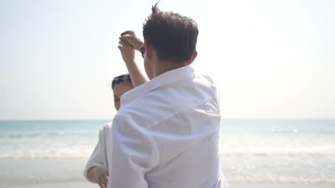 高清慢动作快乐的亚洲高级夫妇男人和女人手牵手，微笑着在海滩上跳舞。退休老年家庭在浪漫婚姻周年旅行中放