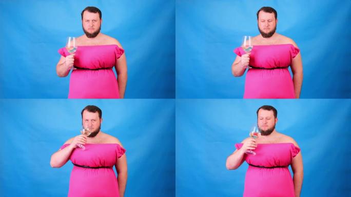 穿着粉红色枕头连衣裙的有趣的大胡子家伙在蓝色背景上喝了一杯酒，很难过。疯狂的检疫。有趣的打扫房子。时