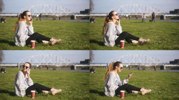 一个女孩坐在公园的草地上谈论视频交流。一个学生，一个自由职业者，微笑着笑。佩珀杯咖啡。旁边有笔记本电