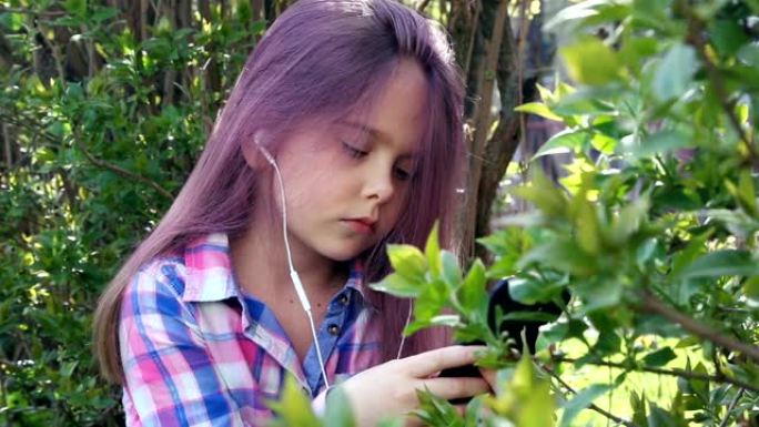 小美丽的女孩在电话上播放，并在树叶灌木和树木的背景上听音乐。