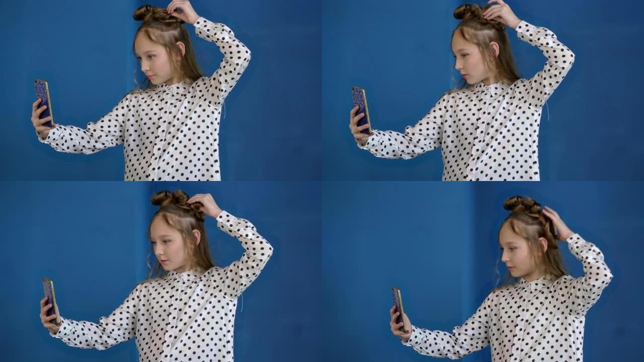 漂亮的少女在蓝色背景上寻找手机相机自拍照片。年轻女子纠正发型，看着像工作室里的镜子一样的智能手机。