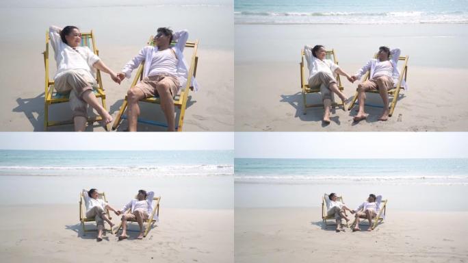 高清慢动作快乐亚洲老年家庭男女躺在沙滩椅上，手牵着手，在沙滩上互相看着。放松退休夫妇在夏天享受浪漫的