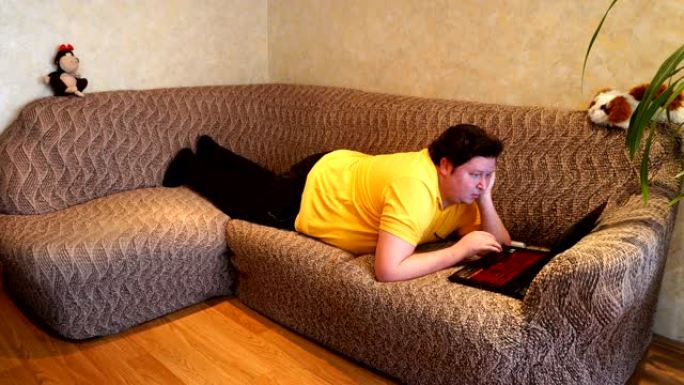 胖子用笔记本电脑躺在客厅的沙发上。年轻人浏览互联网，浏览网络，阅读在线新闻