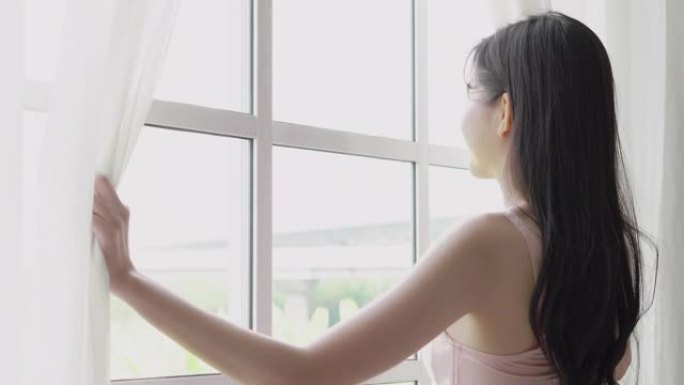 美丽的年轻亚洲女子醒来，早上带着打开的窗帘走到窗前，穿着舒适、性感的内衣女孩微笑着放松，在假期里以快