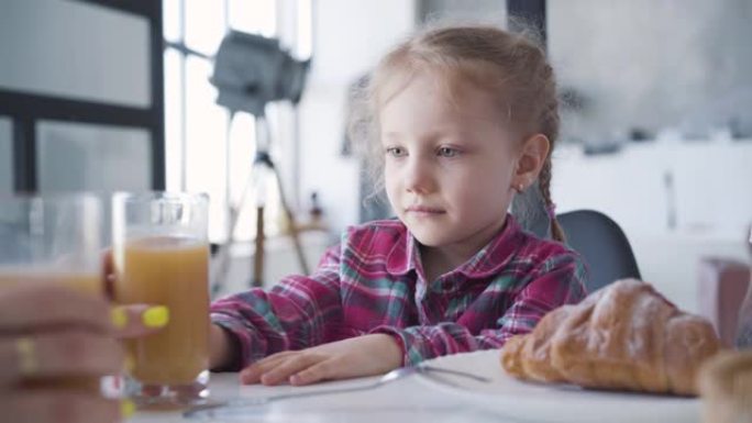 迷人的小女孩早上拒绝喝橙汁的特写肖像。可爱的快乐孩子搬走玻璃，看着妈妈。生活方式，童年，休闲。