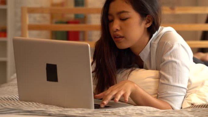 无聊的年轻亚洲女孩躺在床上，在家卧室工作。在线从事计算机工作的女自由职业者。沮丧和强调的表达概念