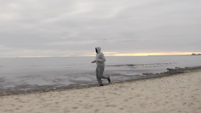 戴着防护口罩的胡须的男子在黄昏时分在海滩上奔跑-多莉射击