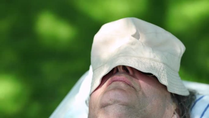 年长的男人在树下被帽子遮住在外面睡着，午睡时特写老人脸午睡