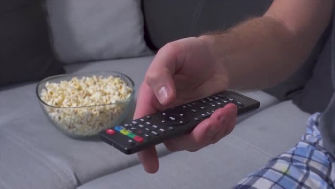 男人手持电视遥控器并按下按钮的手的特写。切换电视频道。沙发上后面有爆米花的碗。
