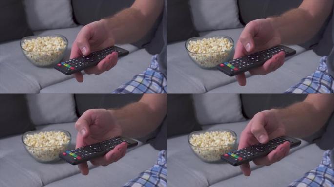 男人手持电视遥控器并按下按钮的手的特写。切换电视频道。沙发上后面有爆米花的碗。