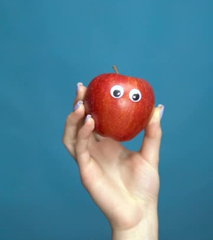 红苹果与眼睛在一个女人的手特写。苹果在蓝色背景上四处张望。垂直视频。